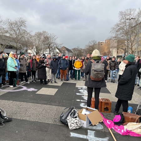 Studierende stehen im Halbkreis vor der Uni Köln und hören einer Rednerin zu.
