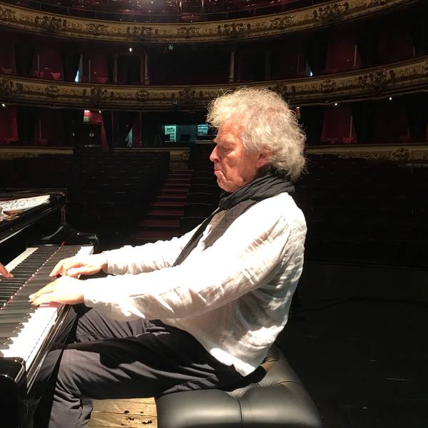 Der Jazz-Musiker Joachim Kühn sitzt in einem leeren Konzertsaal am Klavier. Das Licht ist auf ihn gerichtet und er trägt ein weißes Leinenhemd mit schwarzem Schal.