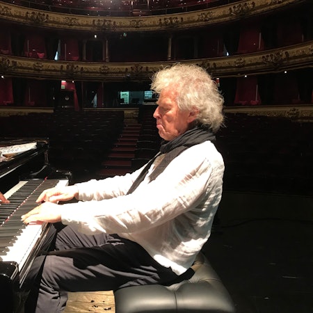 Der Jazz-Musiker Joachim Kühn sitzt in einem leeren Konzertsaal am Klavier. Das Licht ist auf ihn gerichtet und er trägt ein weißes Leinenhemd mit schwarzem Schal.