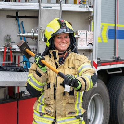 Eine Feuerwehrfrau steht in Einsatzkleidung vor einem Feuerwehrwagen, lächelt in die Kamera und schwingt einen Vorschlaghammer.