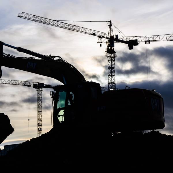 Ein Bagger und einige Baukräne im Hintergrund vor einem bewölkten Himmel