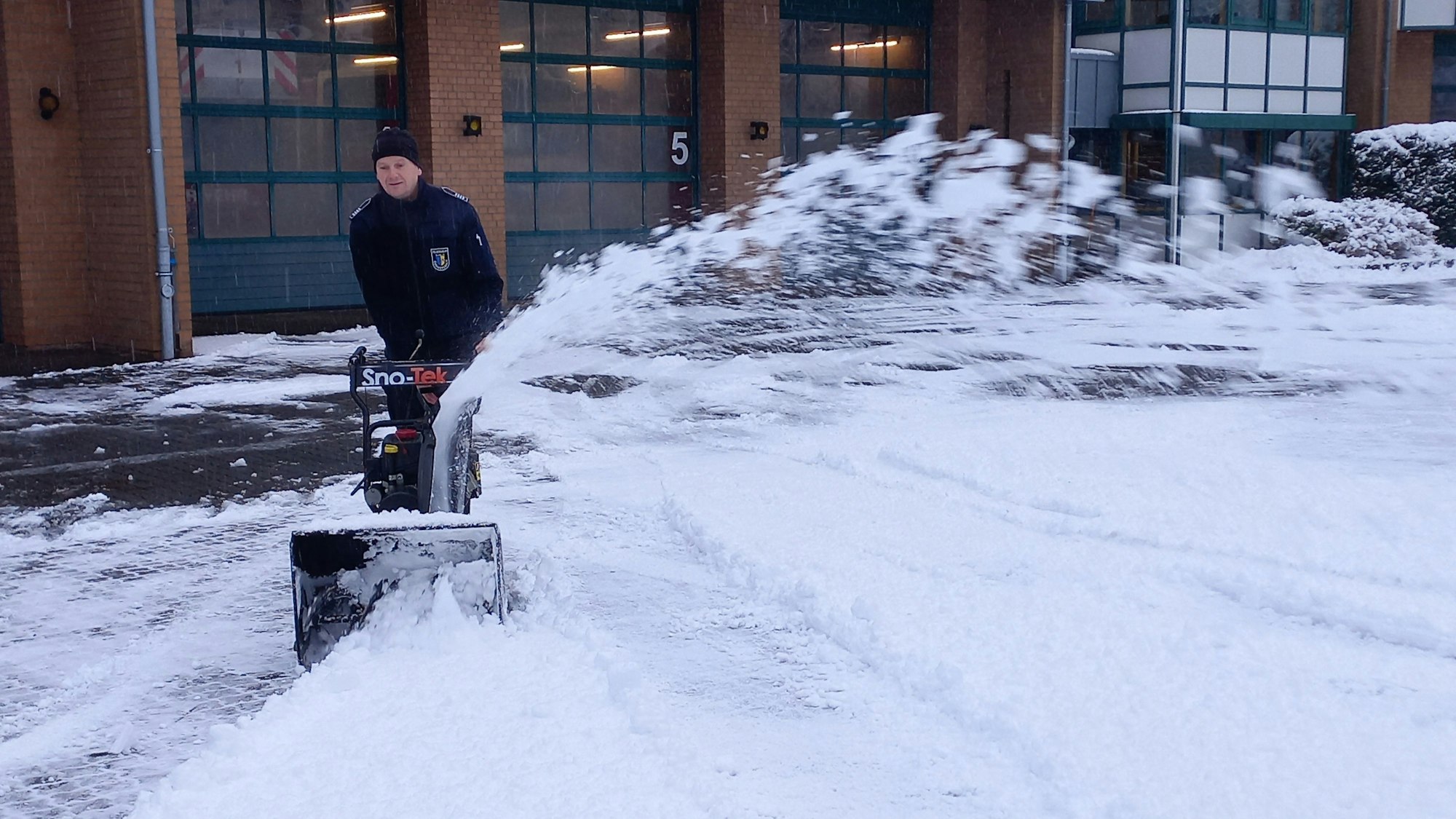 Ein Mann beseitigt mithilfe einer Schneefräse den Schnee auf dem Hof vor einer Feuerwehrwache.