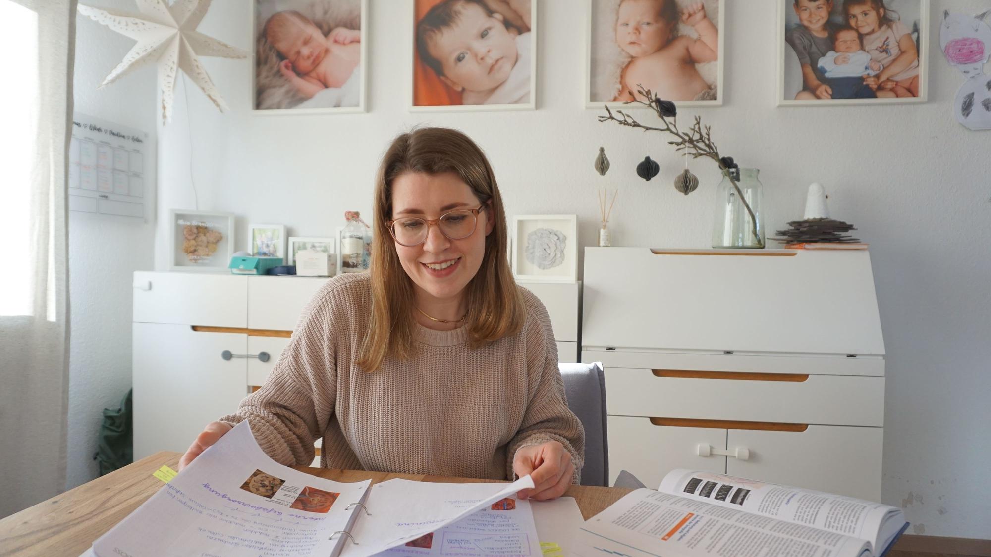 Eine junge Frau sitzt mit Lehrunterlagen am Tisch, im Hintergrund hängen Babyfotos an der Wand.