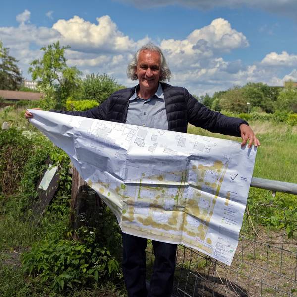 Ein Mann mit längerem grauen Haar steht vor einer abgezäunten Wiese, Er hält einen großen Bauplan in den Händen.