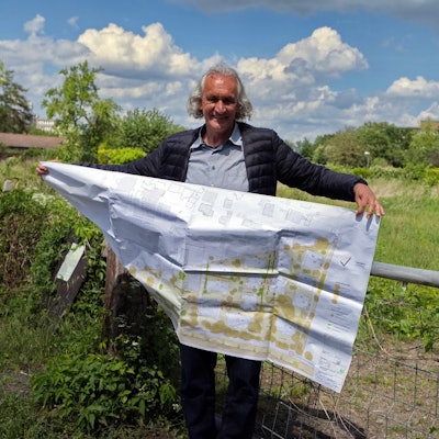 Ein Mann mit längerem grauen Haar steht vor einer abgezäunten Wiese, Er hält einen großen Bauplan in den Händen.