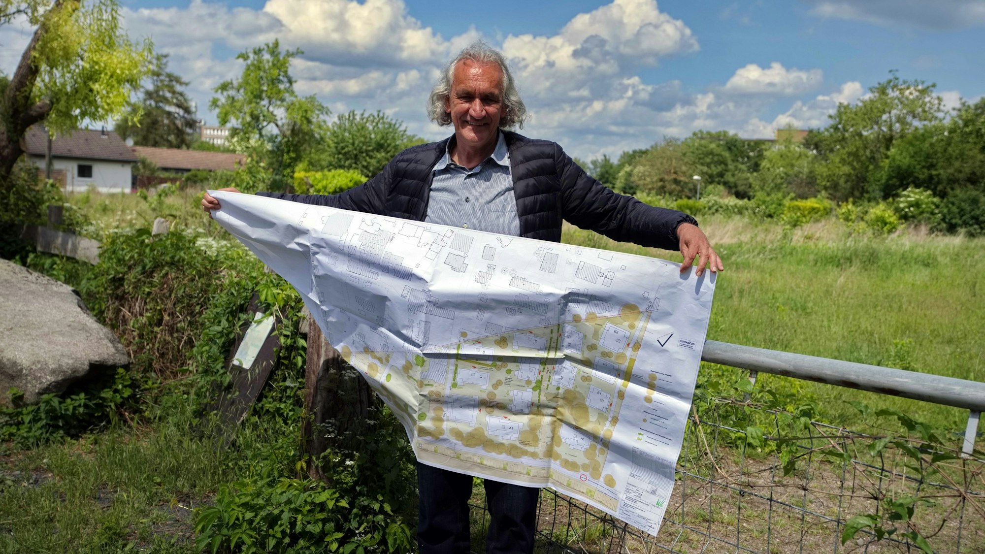 Ein Mann mit längerem grauen Haar steht vor einer abgezäunten Wiese, Er hält einen großen Bauplan in den Händen.