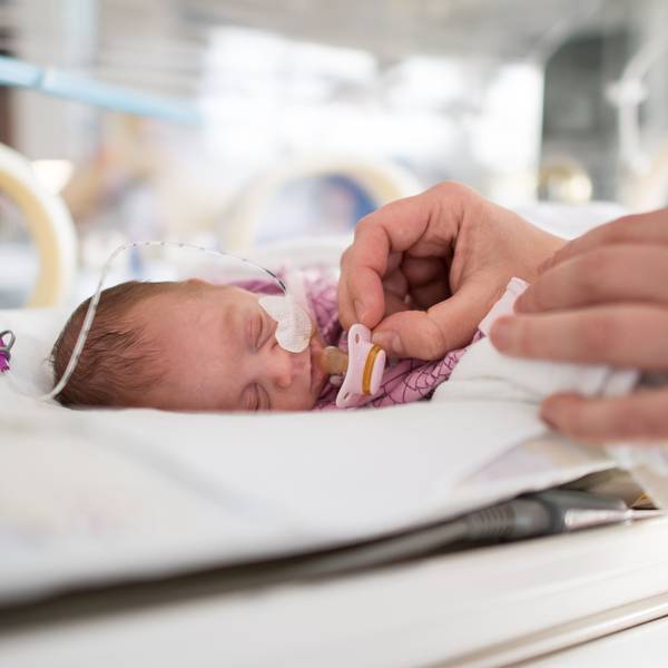 Ein zu früh geborenes Baby liegt im Krankenhaus in einem Inkubator. Eine Pflegekraft hält ihm einen winzigen Schnuller vor den Mund.