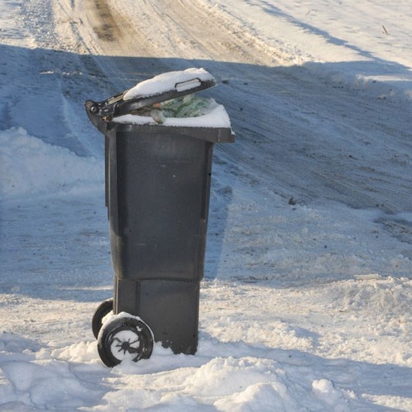 Eine volle Mülltonne steht im Schnee. Im Hintergrund naht ein Räumfahrzeug.