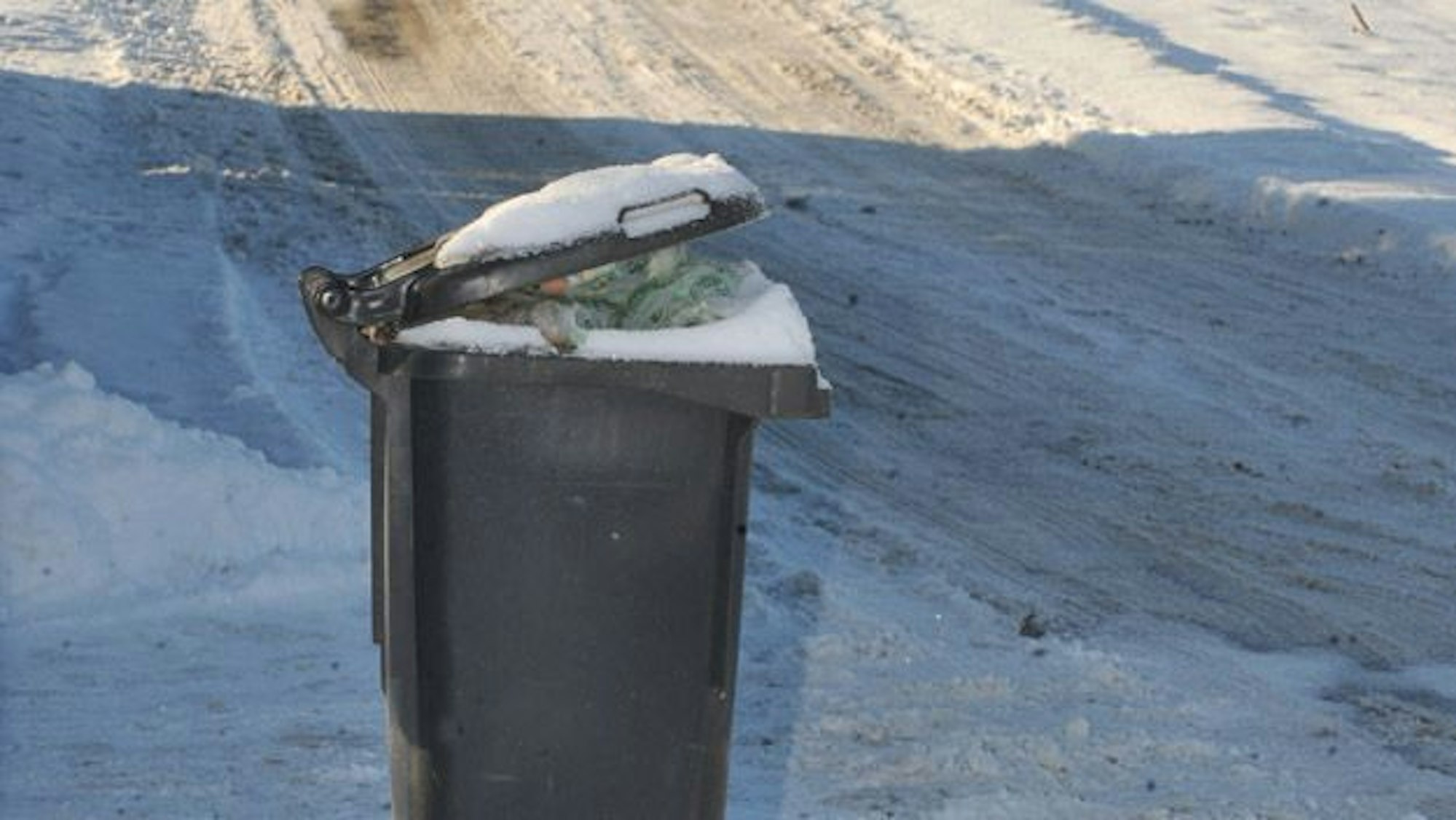 Eine volle Mülltonne steht im Schnee. Im Hintergrund naht ein Räumfahrzeug.