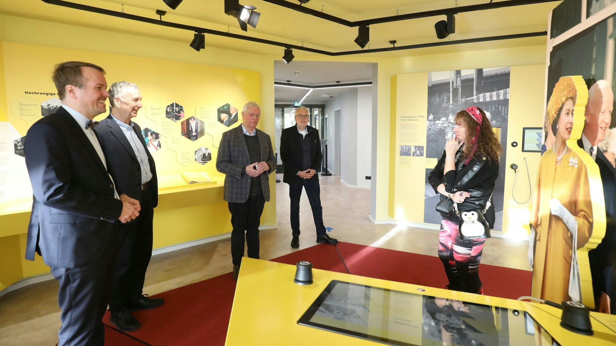 Vier Männer und eine Frau stehen in einem in Gelb gehaltenen Ausstellungsraum des Besucherzentrums für den Petersberg. Rechts am Rand ist lebensgroß ein Bild von Queen Elisabeth II. in einem gelben Kostüm zu sehen.