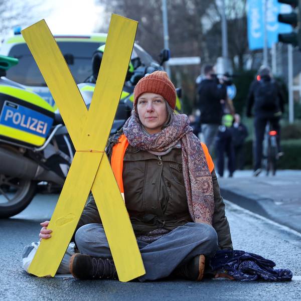 Caroline Schmidt von der Letzten Generation sitzt auf einer Straße im Schneidersitz. Eine Hand ist auf dem Boden angeklebt. In der anderen Hand trägt sie ein gelbes X.