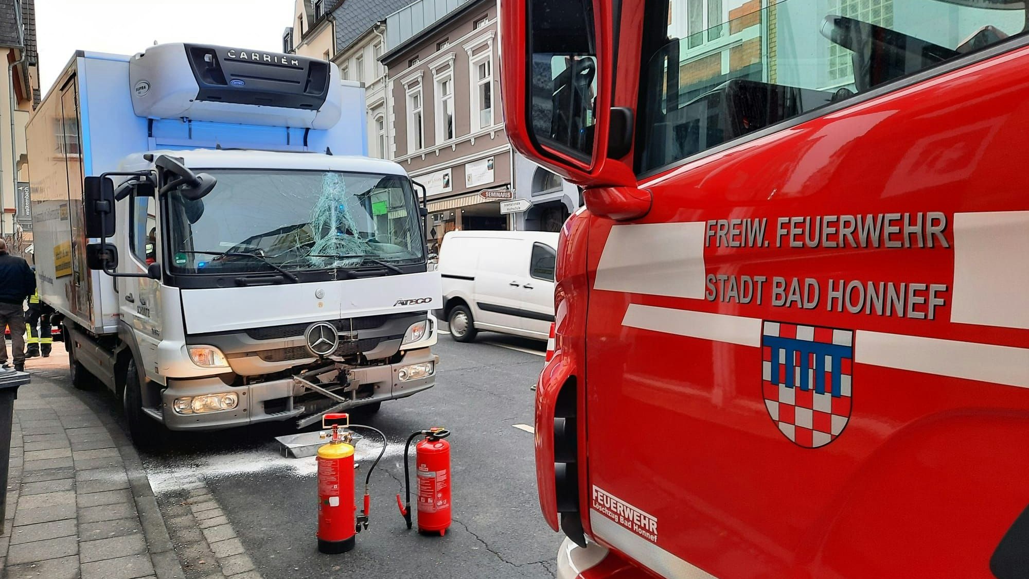 Ein rotes Feuerwehrfahrzeug steht vor dem Lastwagen, dessen Frontscheibe durch den Unfall zum Teil eingedrückt wurde. Zwei Feuerwehrlöscher stehen sicherheitshalber vor dem Unfallfahrzeug.
