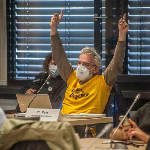 Benedikt Rees sitzt in einem gelben Shirt vor einem geöffneten Tablet und meldet sich mit beiden Händen in die Höhe gestreckt. Er träckte einen Mund-Nasen-Schutz.