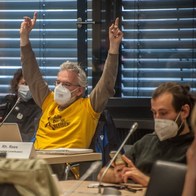 Benedikt Rees sitzt in einem gelben Shirt vor einem geöffneten Tablet und meldet sich mit beiden Händen in die Höhe gestreckt. Er träckte einen Mund-Nasen-Schutz.