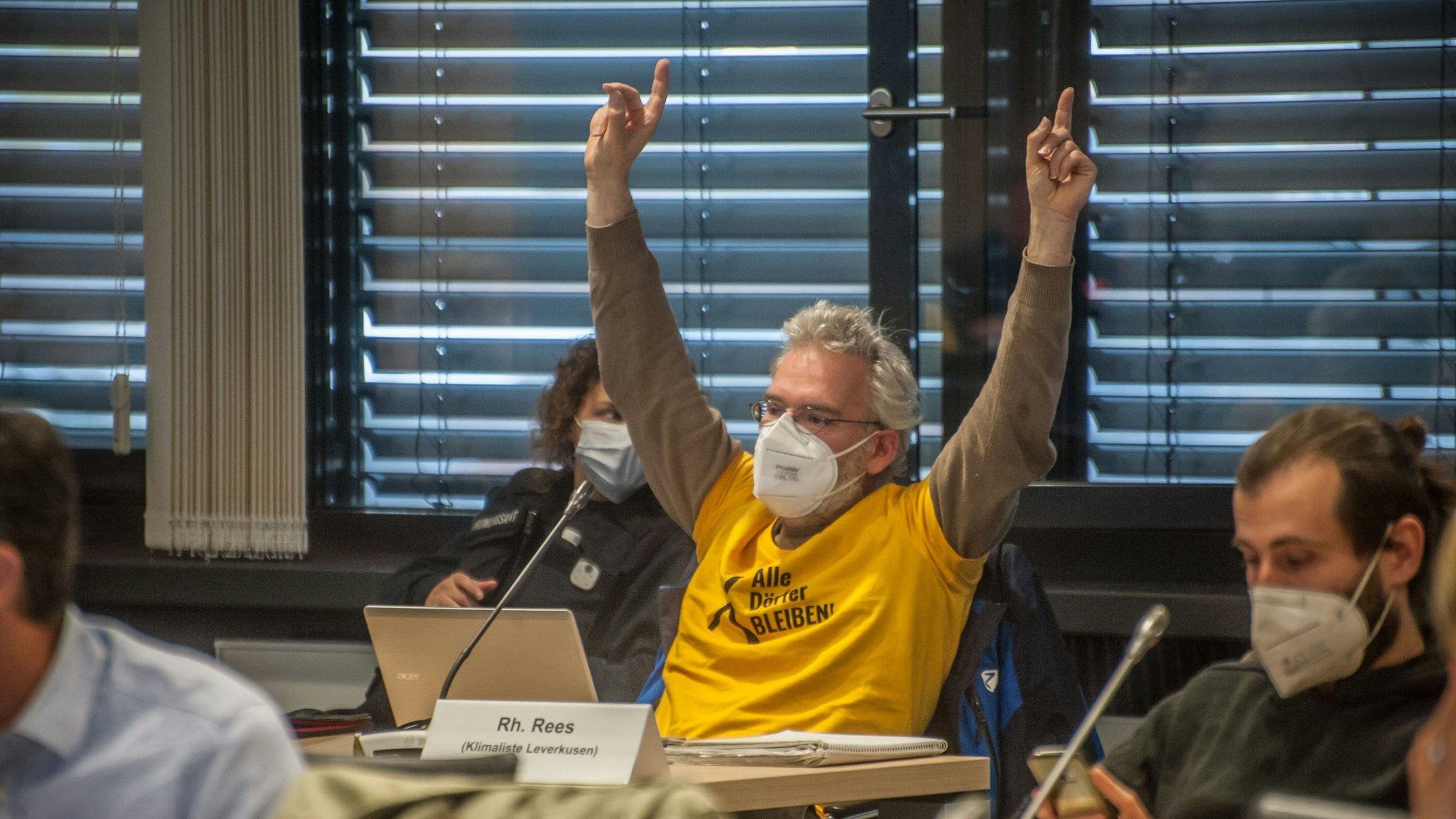 Benedikt Rees sitzt in einem gelben Shirt vor einem geöffneten Tablet und meldet sich mit beiden Händen in die Höhe gestreckt. Er träckte einen Mund-Nasen-Schutz.
