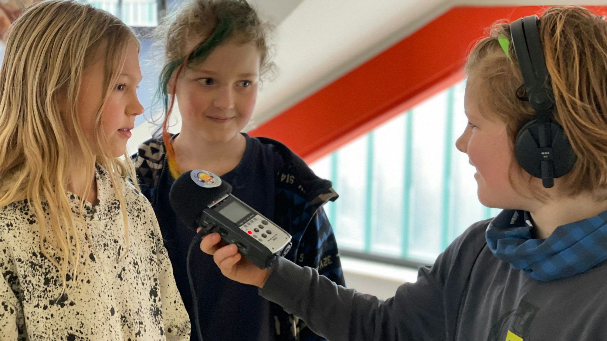 Ein Kind trägt Kopfhörer und hält seinen Mitschülerinnen ein Mikrofon hin.