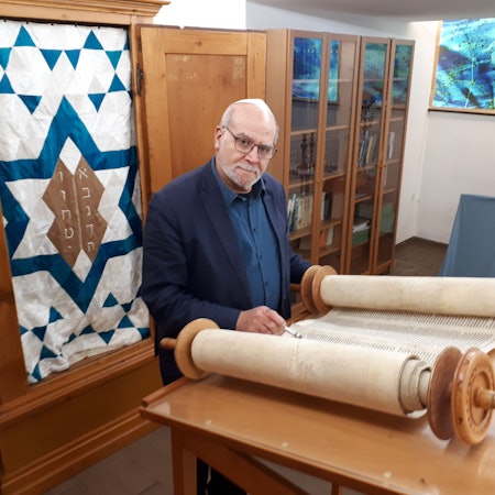 Rafi Rothenberg steht vor einer Fahne, die in eine Schrankwand integriert ist. Im Hintergrund rechts zwei Leuchter.