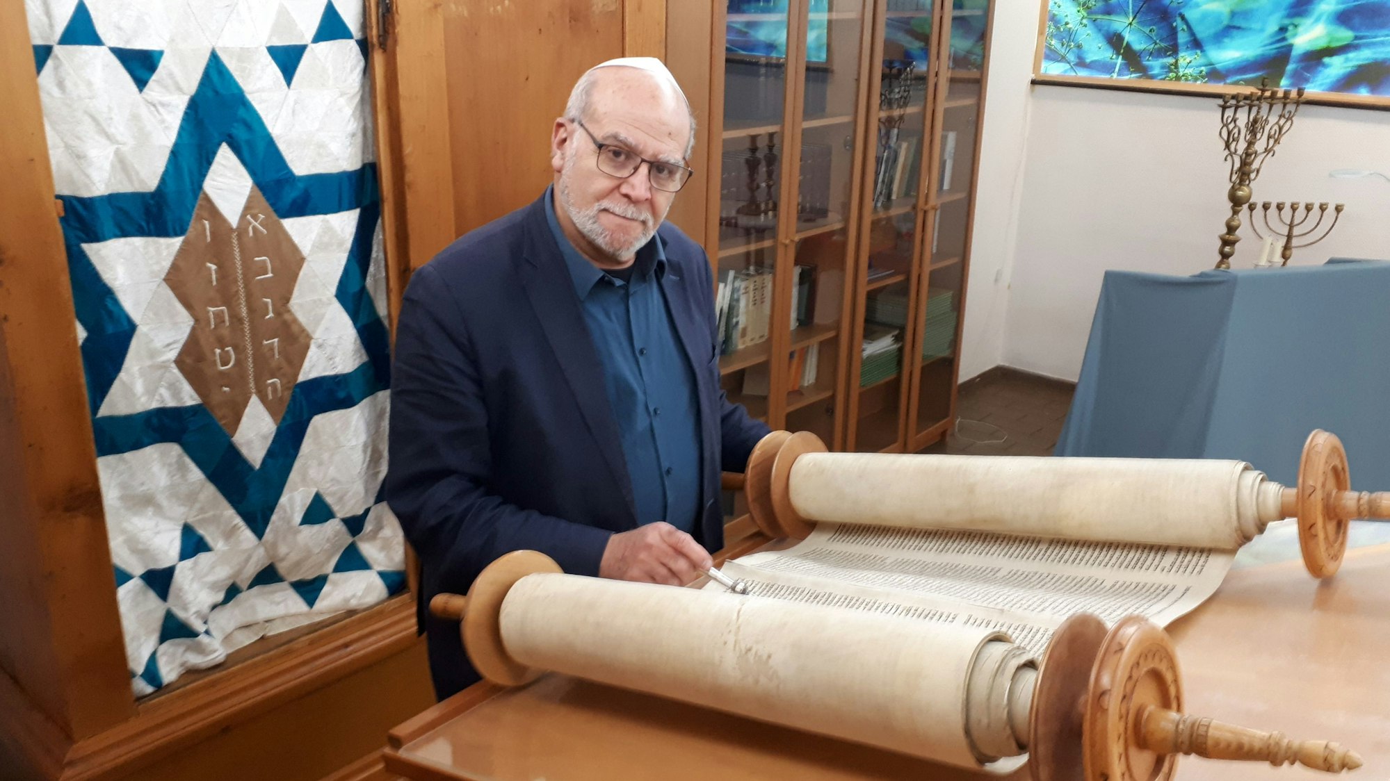 Rafi Rothenberg steht vor einer Fahne, die in eine Schrankwand integriert ist. Im Hintergrund rechts zwei Leuchter.