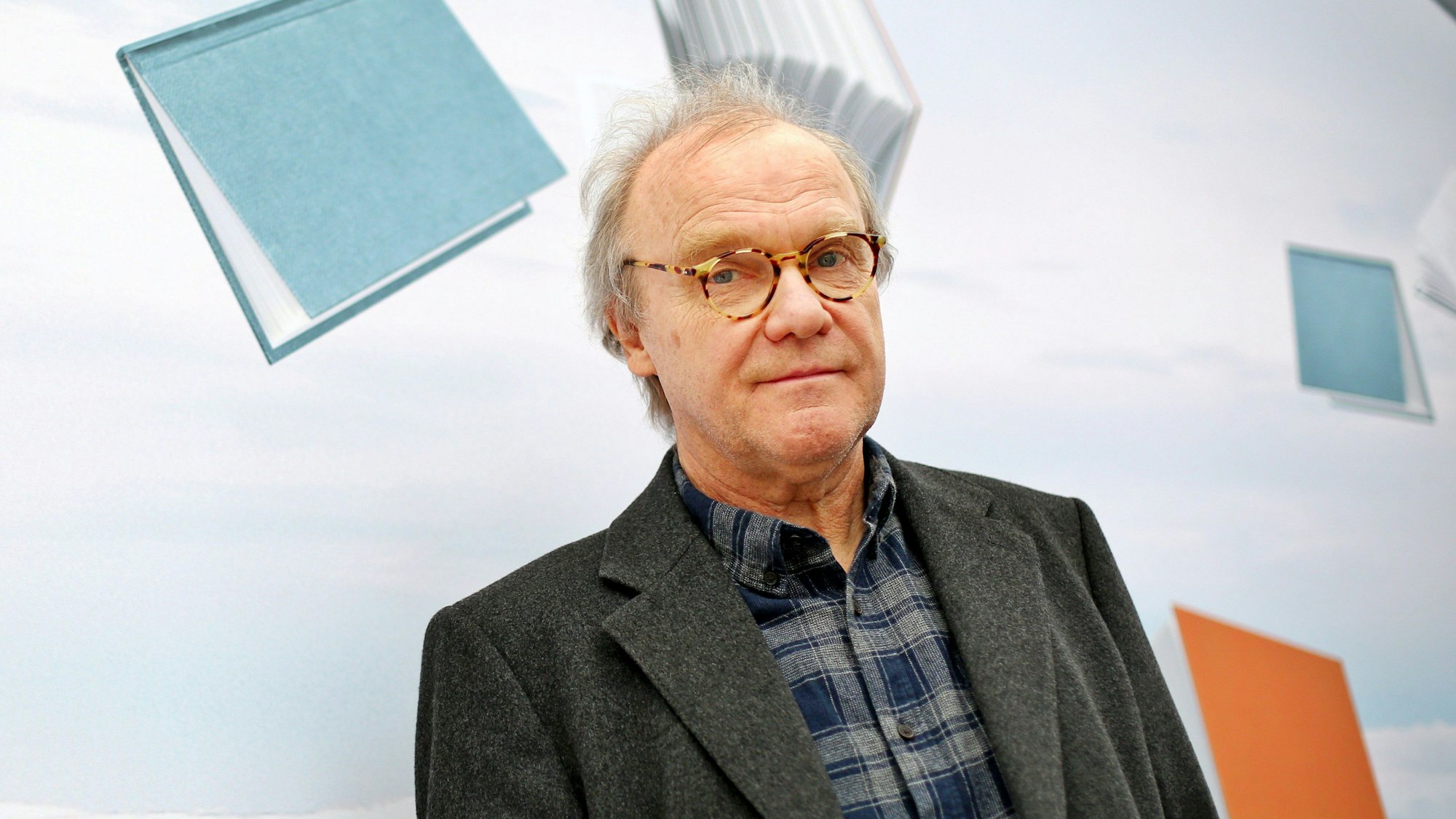 Der österreichische Autor Michael Köhlmeier steht in einem dunklen Jackett und einem karierten Hemd vor einer mit Büchern bedruckten Wand.