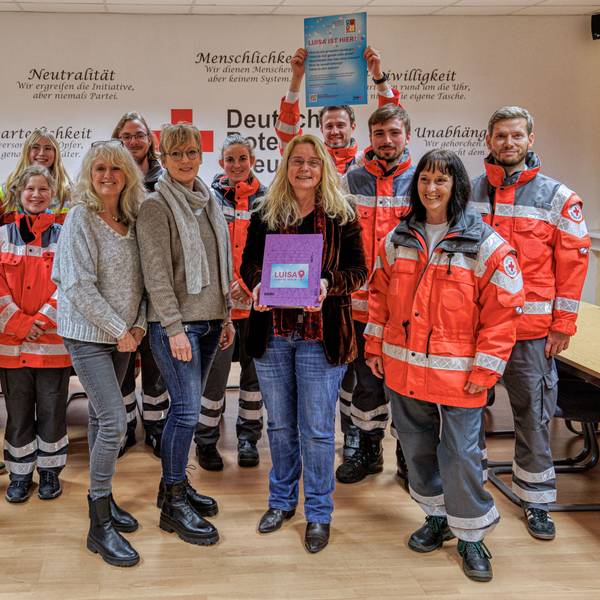 Das Team des DRK steht in neonroter Arbeitskleidung in einem Büro und hält Infomaterial in der Hand.