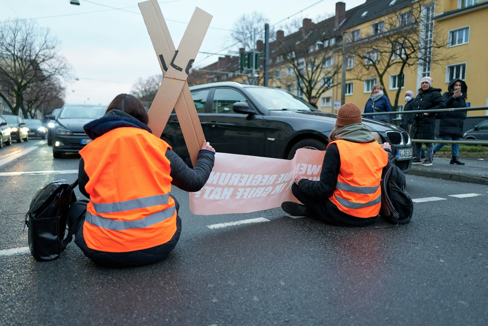 Aktivisten der „Letzten Generation“ kleben sich auf der Fahrbahn fest. Einige Autofahrer versuchen, an den Aktivisten vorbeizukommen.