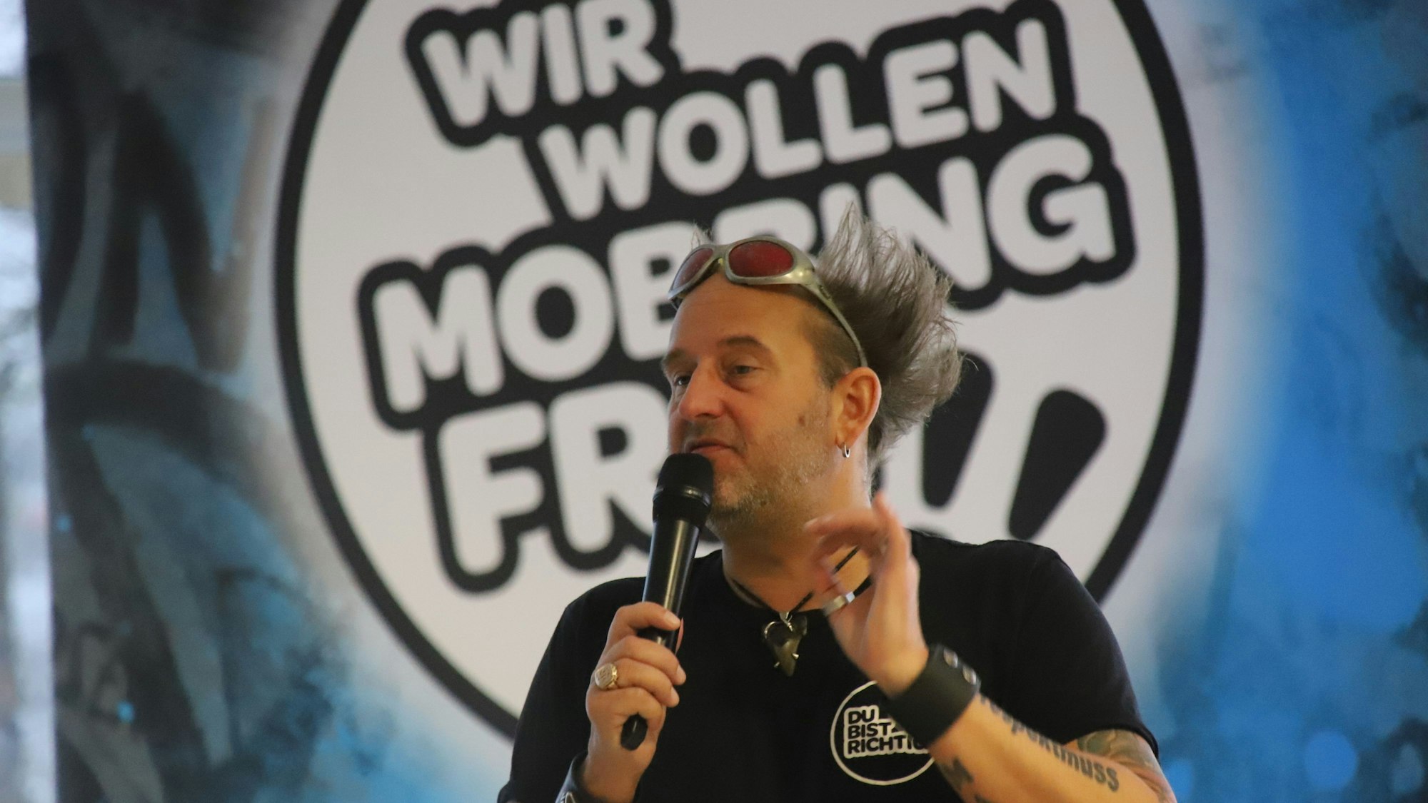 Ein Mann mit einem Mikrofon in der Hand gestikuliert. Er trägt ein T-Shirt und eine Sonnenbrille mit roten Gläsern oben auf der Stirn. Seine Haare stehen borstig ab, er steht vor einem runden Schild mit der Aufschrift „Wir wollen mobbingfrei“.