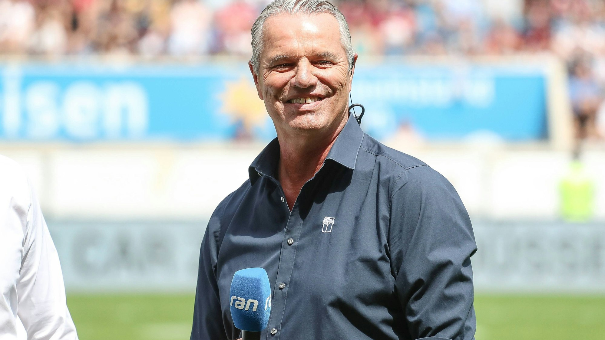 Jan Stecker steht mit einem blauen Mikrofon in der Hand in einem Stadion.