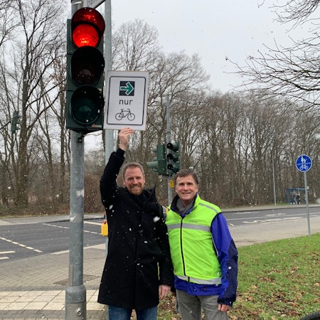 An einer roten Ampel stehen zwei Männer, einer hält ein Schild mit einem grünen Pfeil und dem Symbol eines Fahrrades in die Höhe.