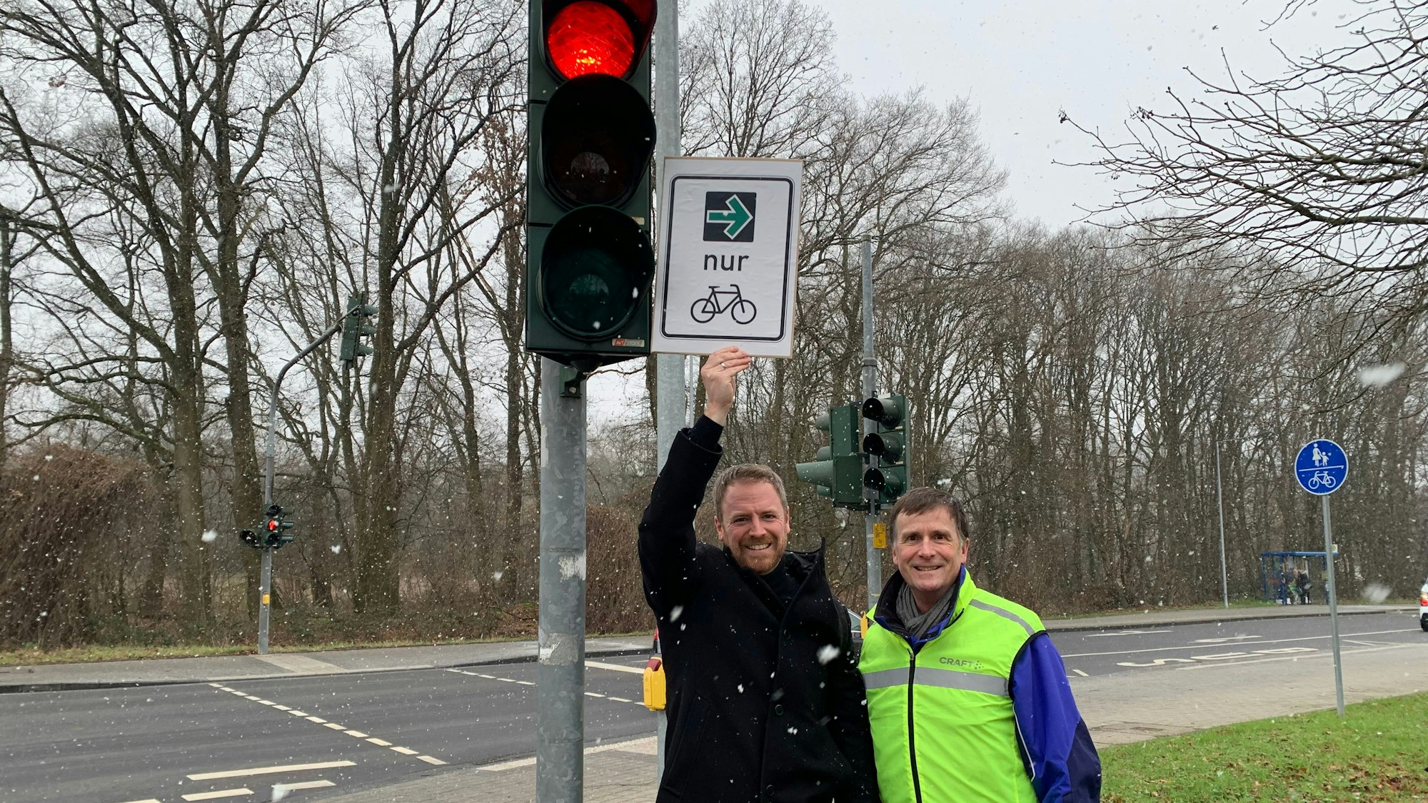 An einer roten Ampel stehen zwei Männer, einer hält ein Schild mit einem grünen Pfeil und dem Symbol eines Fahrrades in die Höhe.