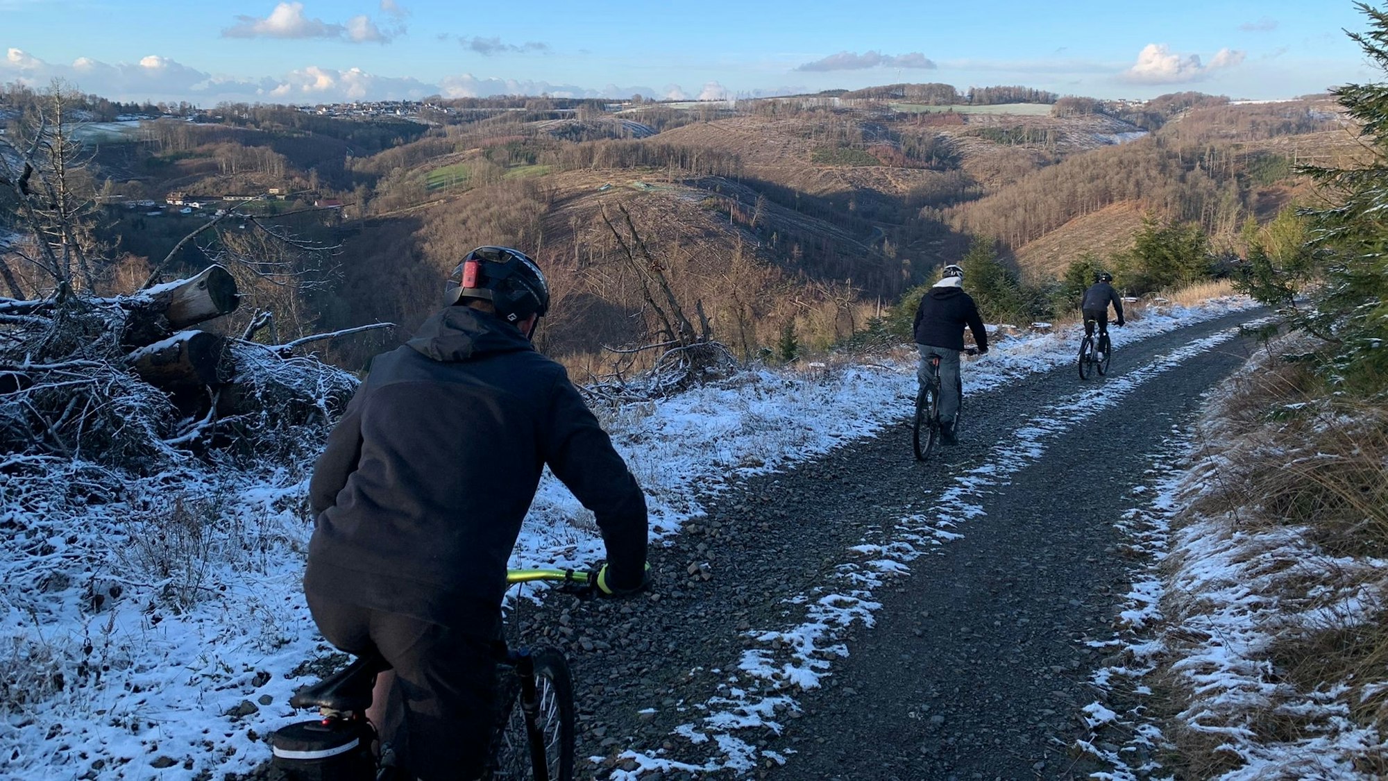 Drei Fahrradfahrer sind von hinten zu sehen, wie sie über einen Schotterweg mit ihren Mountainbikes einen Berg runter fahren.