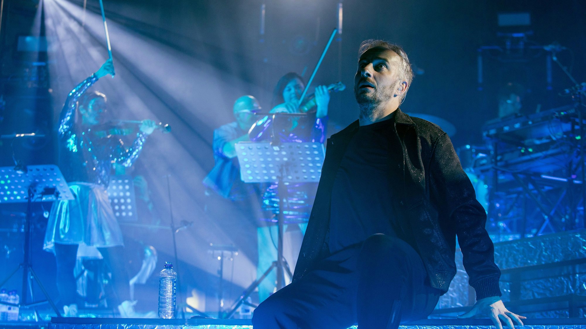 Jan Böhmermann sitzt auf der Bühne des Palladium, während hinter ihm die Streicherinnen vom Rundfunk-Tanzorchester spielen.