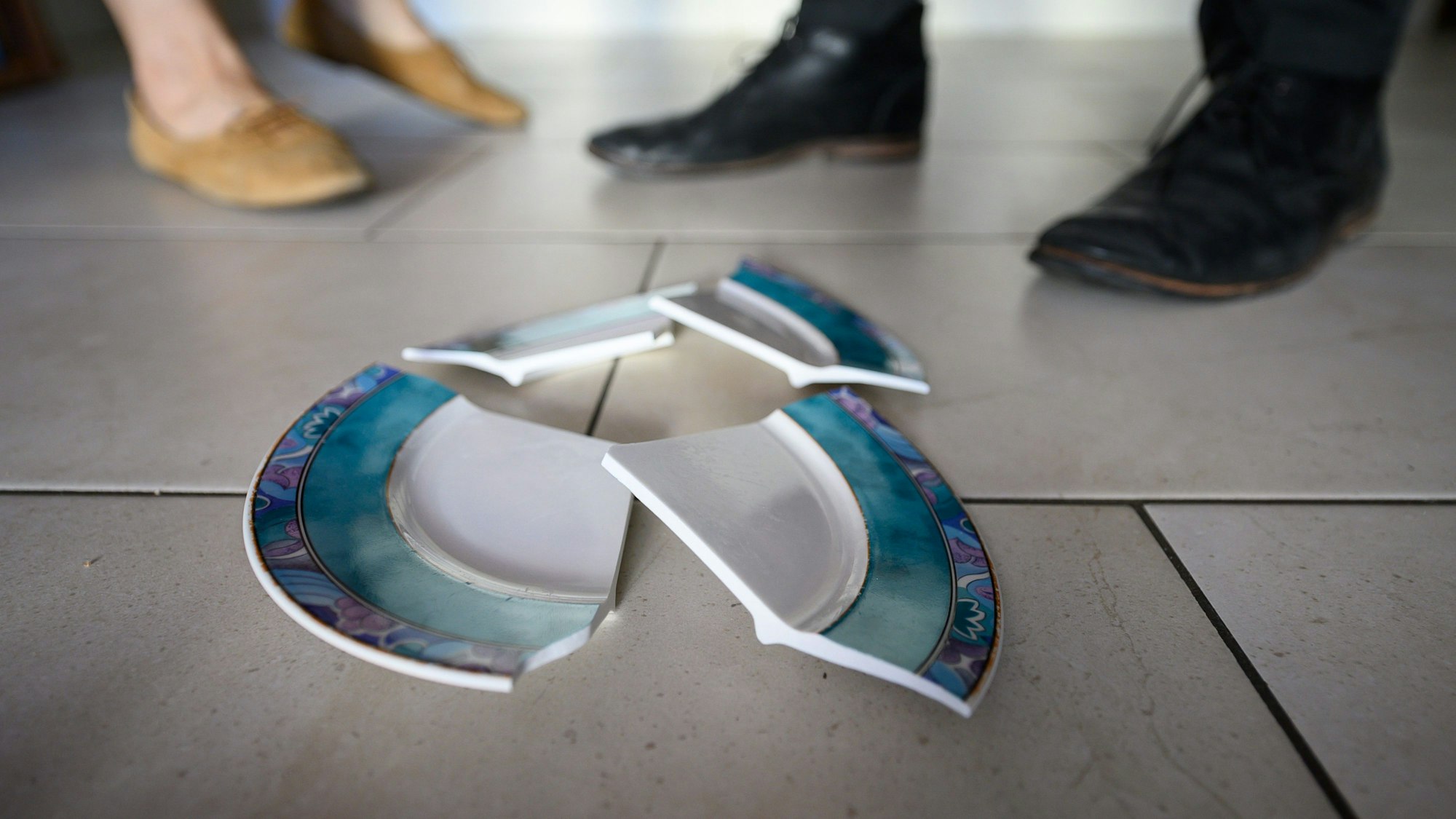 Ein zerbrochener Teller liegt auf dem Boden. Im Hintergrund stehen ein Mann und eine Frau.