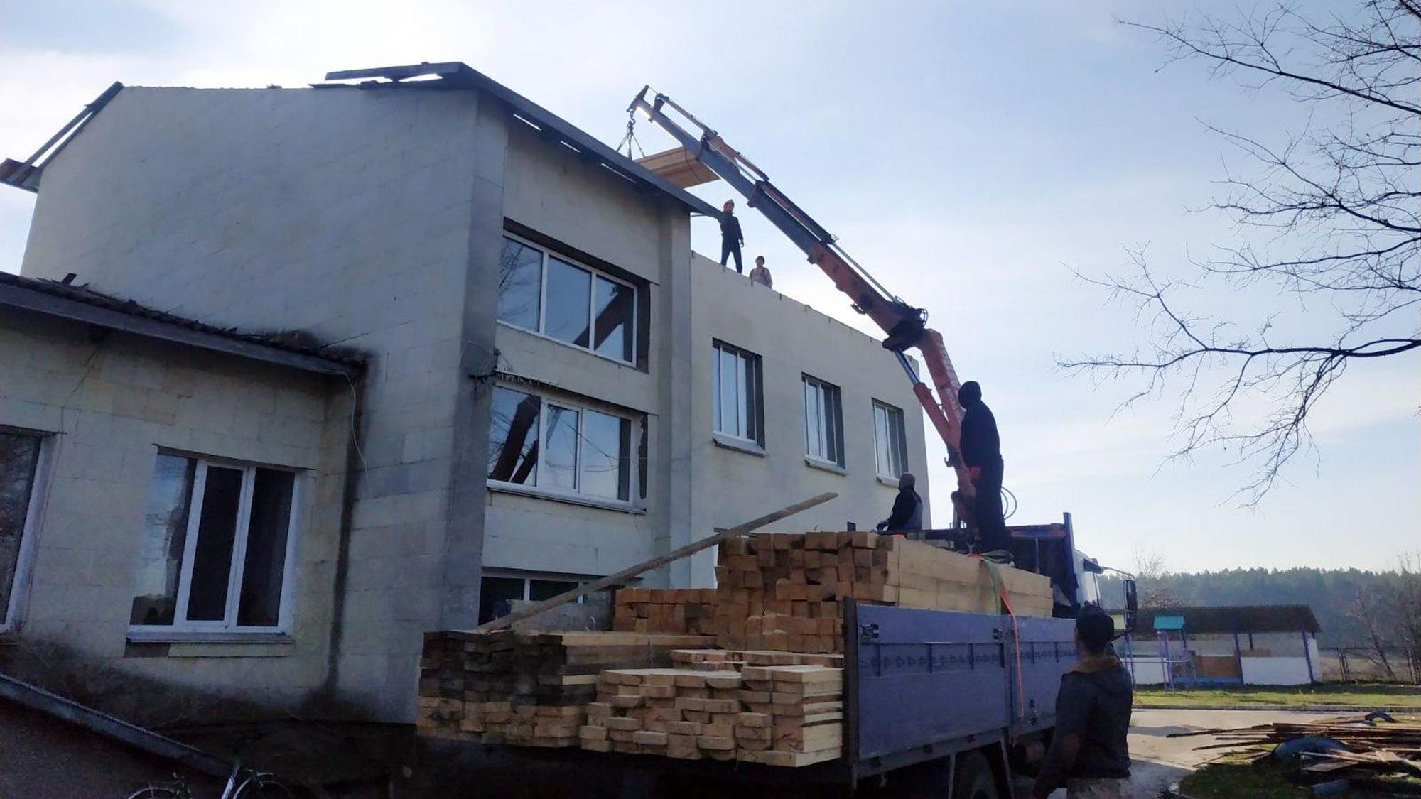 Ein Kran transportiert Holzbalken von einem Lastwagen auf das Obergeschoss eines Gebäudes ohne Dach.
