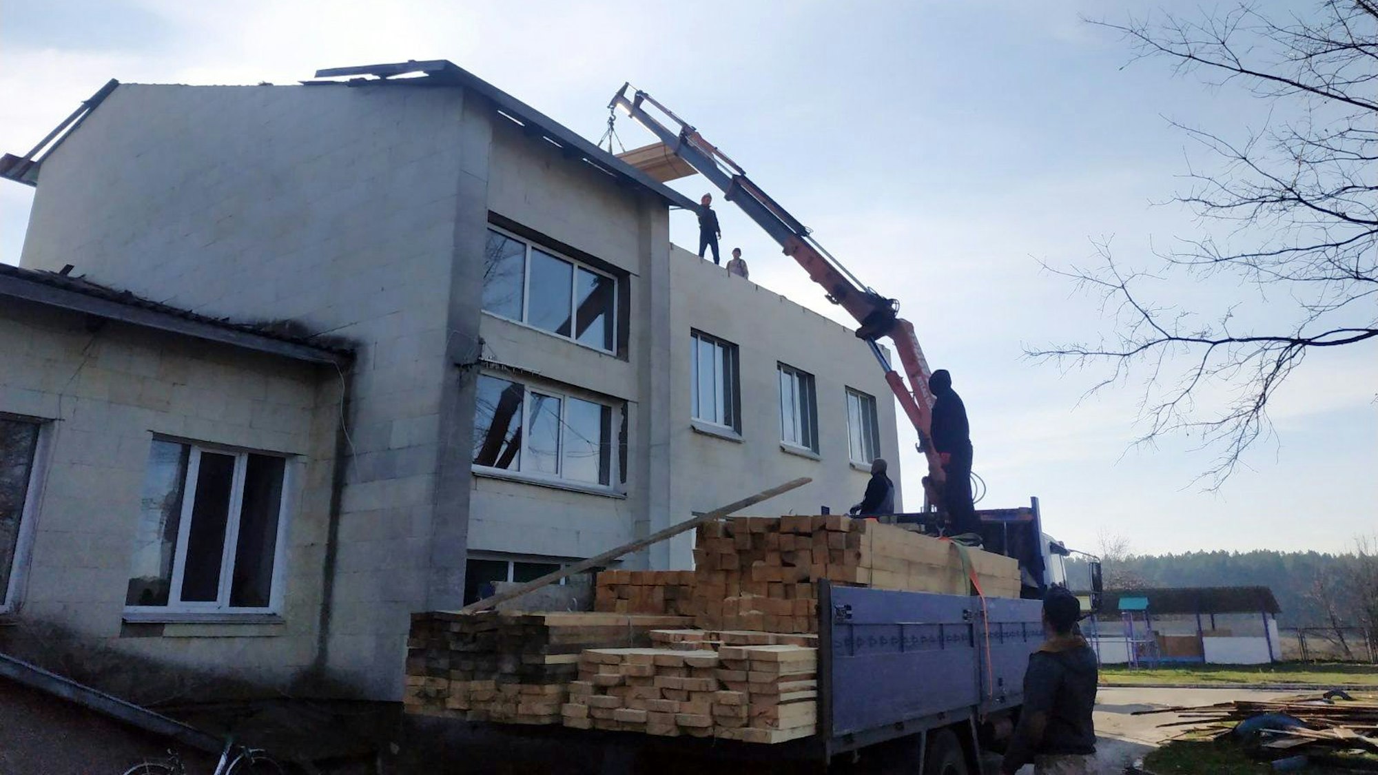 Ein Kran transportiert Holzbalken von einem Lastwagen auf das Obergeschoss eines Gebäudes ohne Dach.