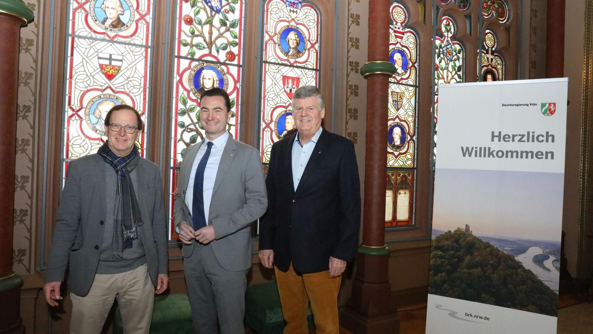 Drei Männer stehen vor den großen Buntglasfenstern in der Kunsthalle von Schloss Drachenburg, Ein Aufsteller am rechten Rand zeigt die Ruine des Drachenfels' mit dem Rhein.