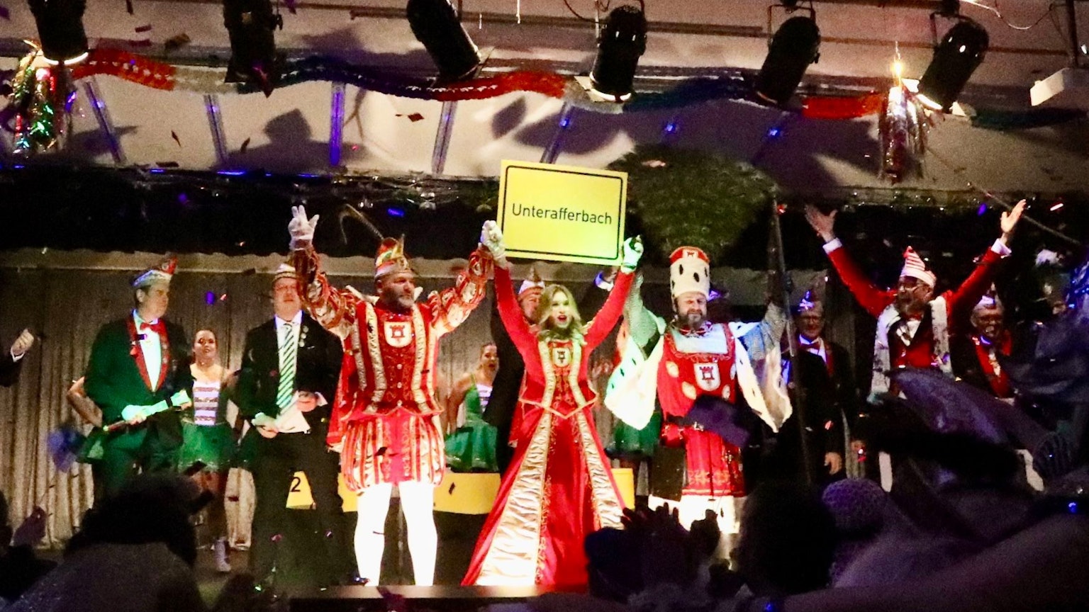 Das Porzer Dreigestirn in Unterafferbach auf der Bühne. Sie halten ein Ortsschild von Unterafferbach hoch.