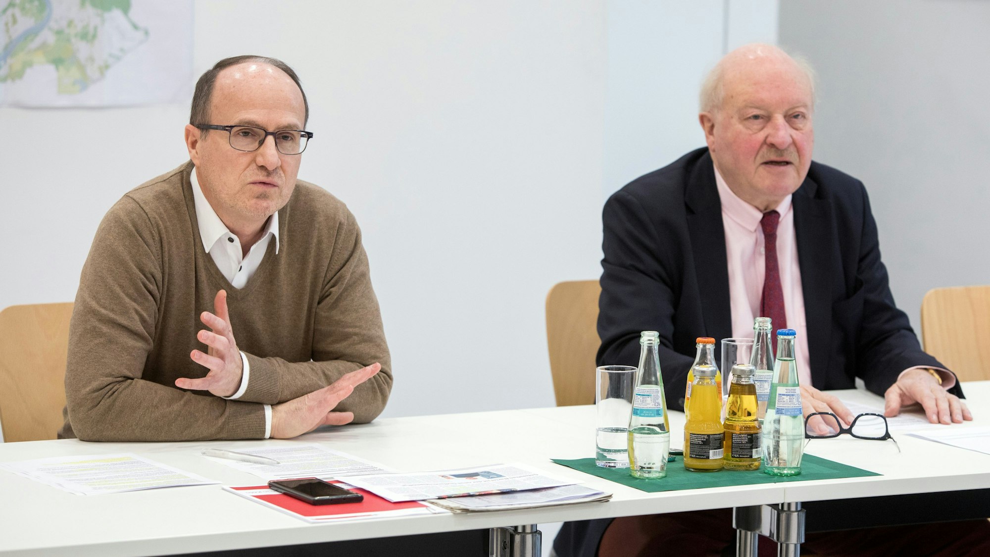 Ein mittelalter Mann mit Hemd, Pullover und Brille und ein älterer Mann mit Sakko, Hemd und Krawatte sitzen an einem Tisch nebeneinander