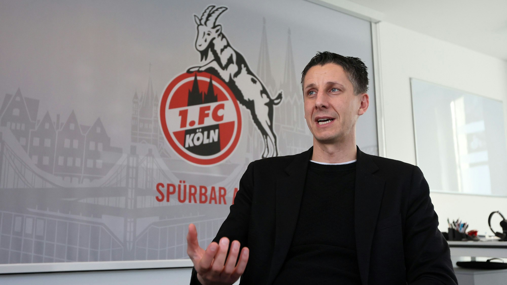 Christian Keller, Geschäftsführer des 1. FC Köln, gestikuliert vor einem FC-Plakat mit Geißbock