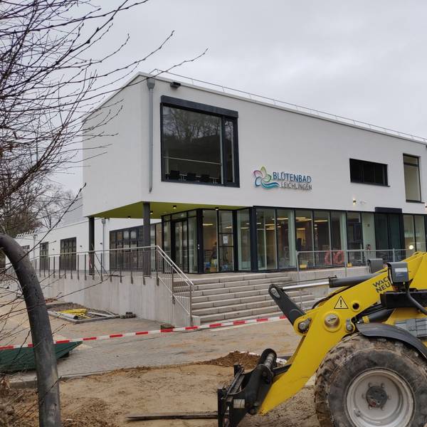 Ein Bagger steht vor dem Eingang des abgesperrten Hallenbad-Neubaus. Das neue blau-grüne Blütenbad-Logo hängt an der weißen Fassade.