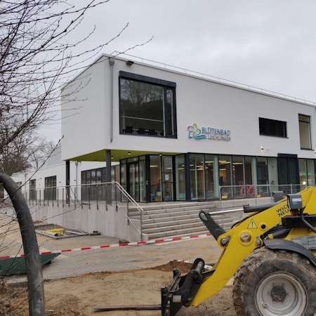 Ein Bagger steht vor dem Eingang des abgesperrten Hallenbad-Neubaus. Das neue blau-grüne Blütenbad-Logo hängt an der weißen Fassade.