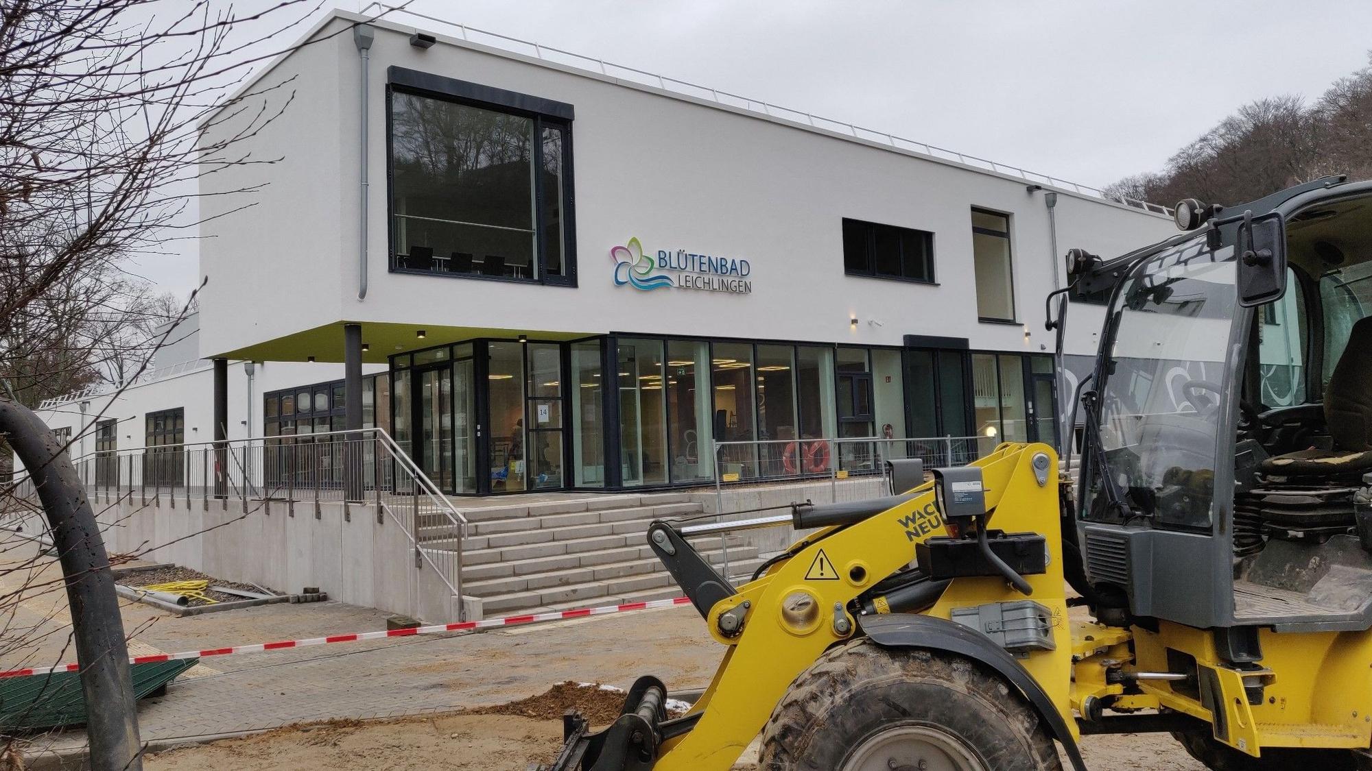 Ein Bagger steht vor dem Eingang des abgesperrten Hallenbad-Neubaus. Das neue blau-grüne Blütenbad-Logo hängt an der weißen Fassade.
