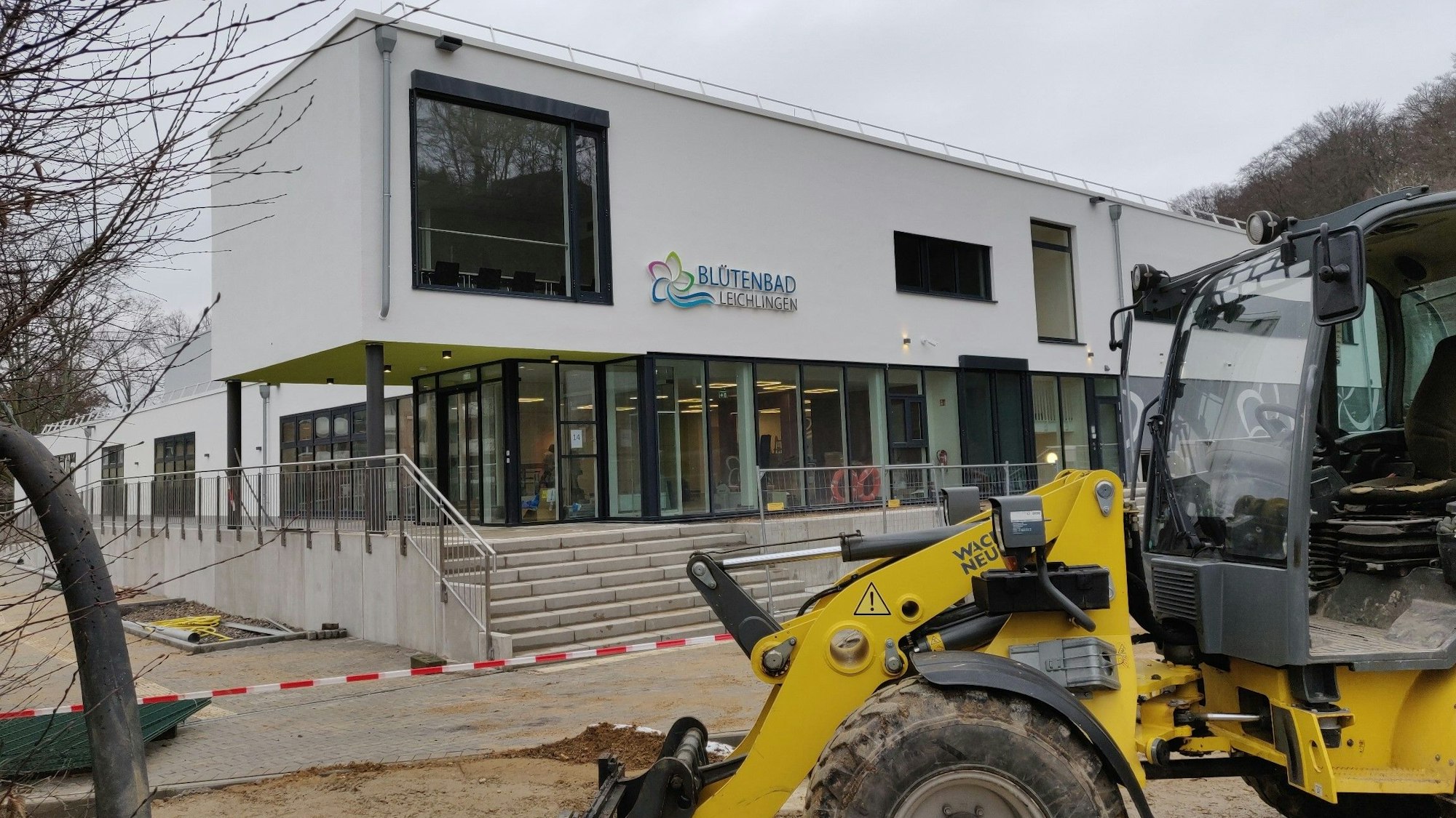 Ein Bagger steht vor dem Eingang des abgesperrten Hallenbad-Neubaus. Das neue blau-grüne Blütenbad-Logo hängt an der weißen Fassade.