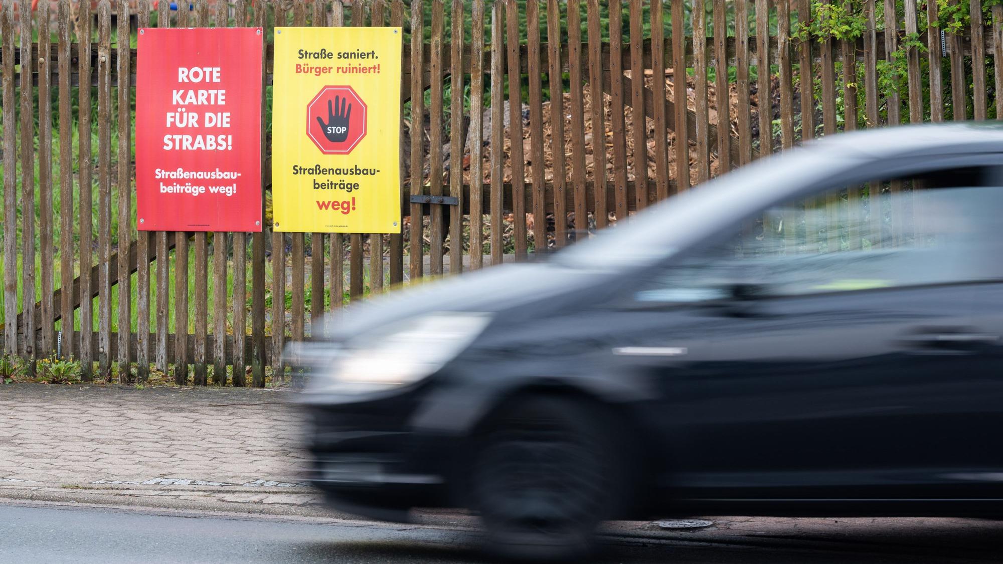 Schilder mit der Aufschrift „Rote Karte für die Strabs! Straßenbaubeiträge weg!“sowie „Straße saniert, Bürger ruiniert“ hängen am Zaun an einem Grundstück.