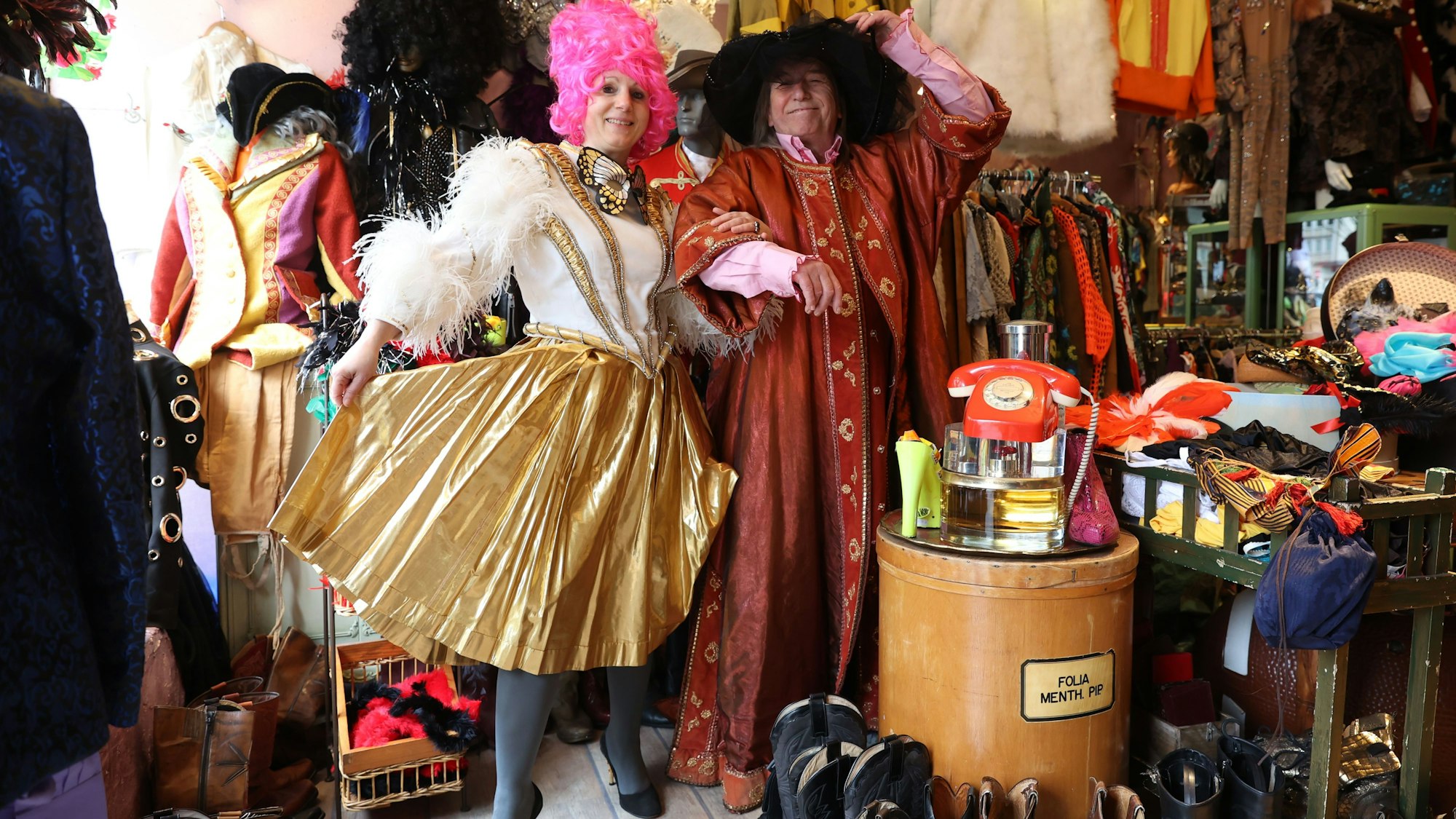 Eine Fau in einem gold-weißen Fransen-Dress und ein Mann in einer rot-goldenen Robe schunkeln in dem dem Second-Hand-Geschäft.