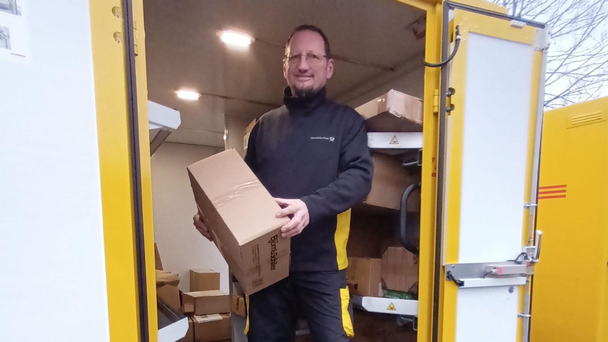 Ein Mann steht in einem geöffneten Zustellfahrzeug und hält ein Paket in den Händen. Hinter ihm liegen etliche Päckchen und Pakete in Regalen.