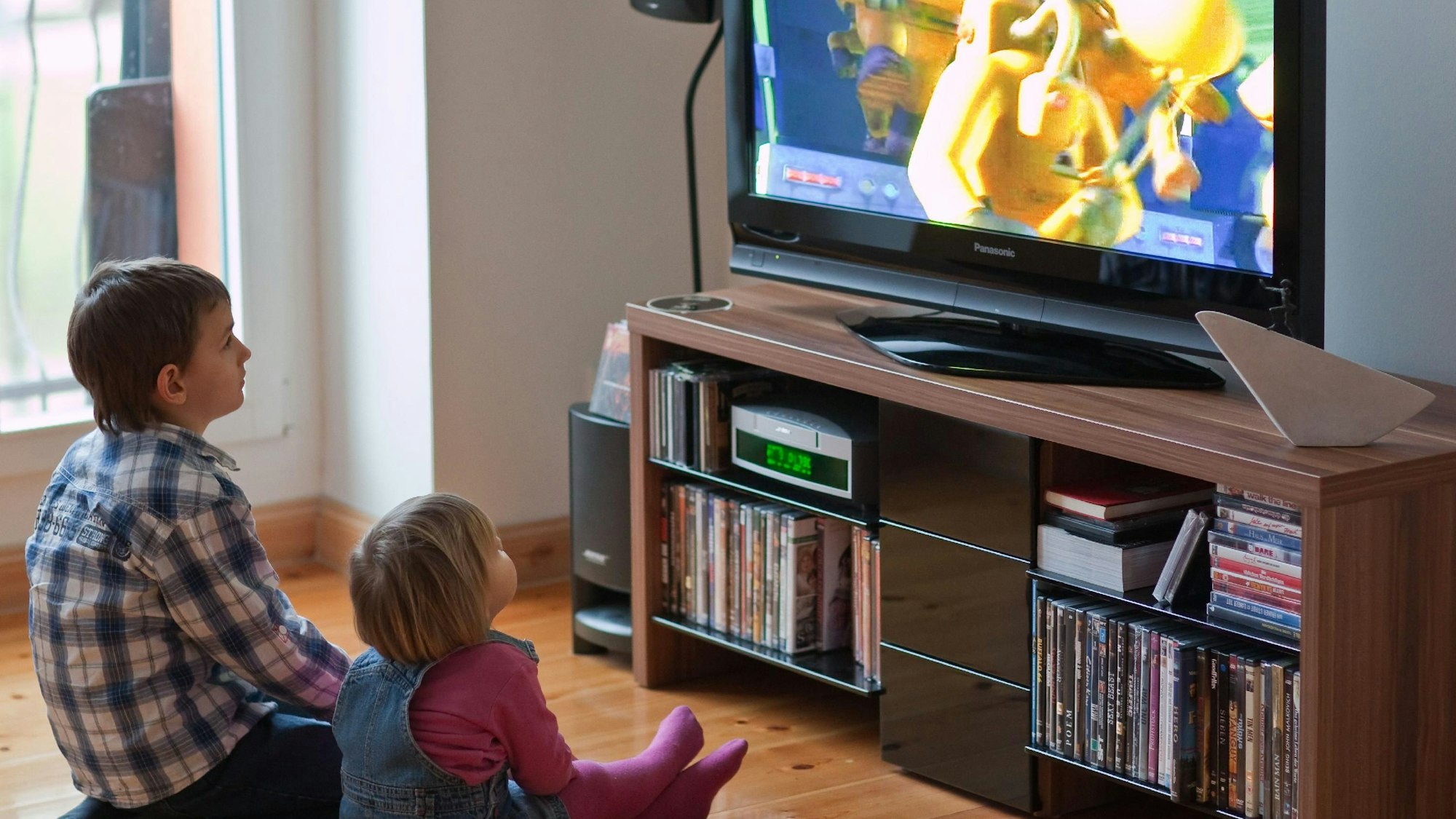 Zwei Kinder sitzen vor einem laufenden Fernseher auf dem Boden.