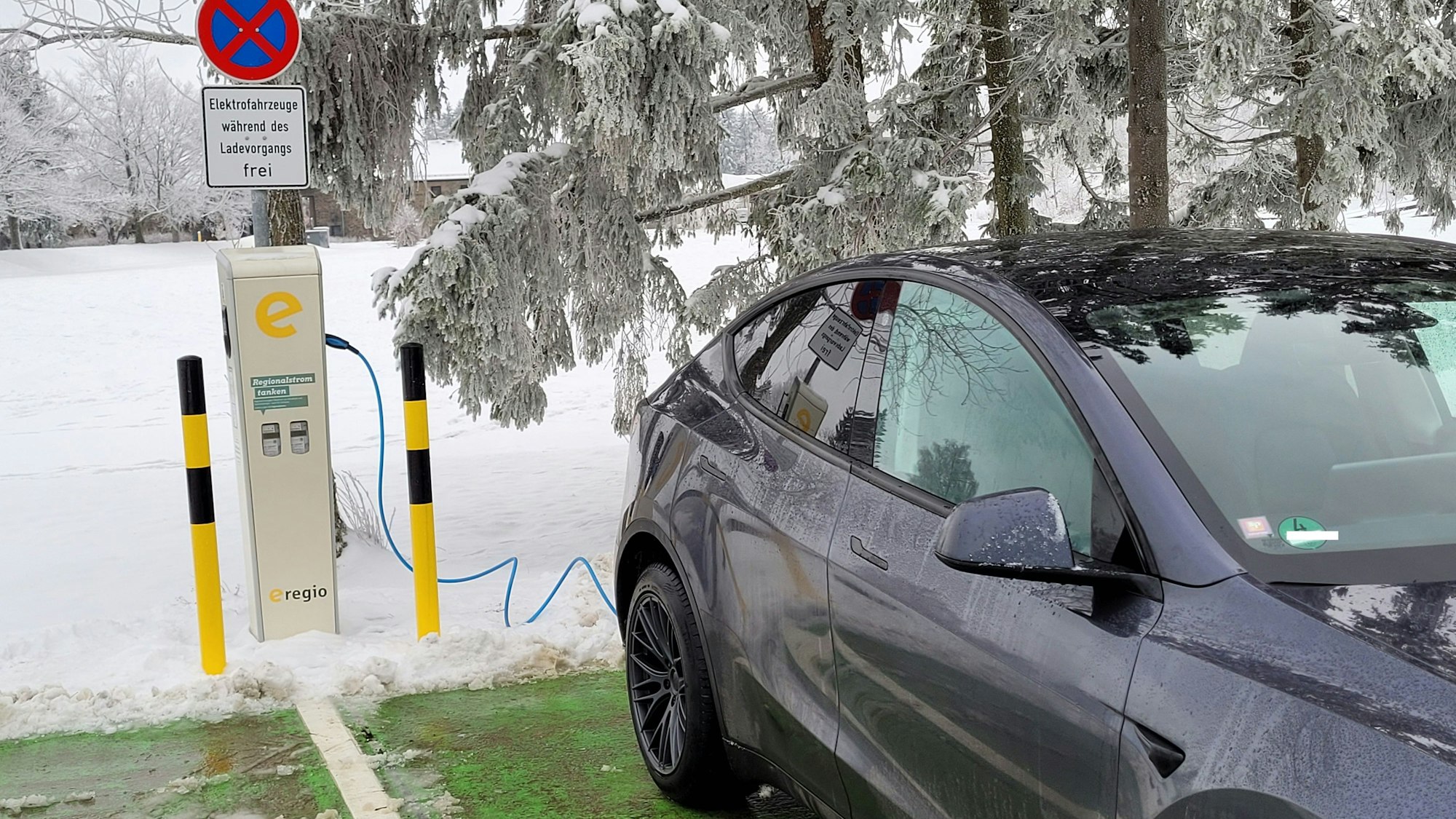 Ein Elektroauto steht an einer Ladestation, im Hintergrund ein Baum und eine Wiese, die mit Schnee bedeckt sind.