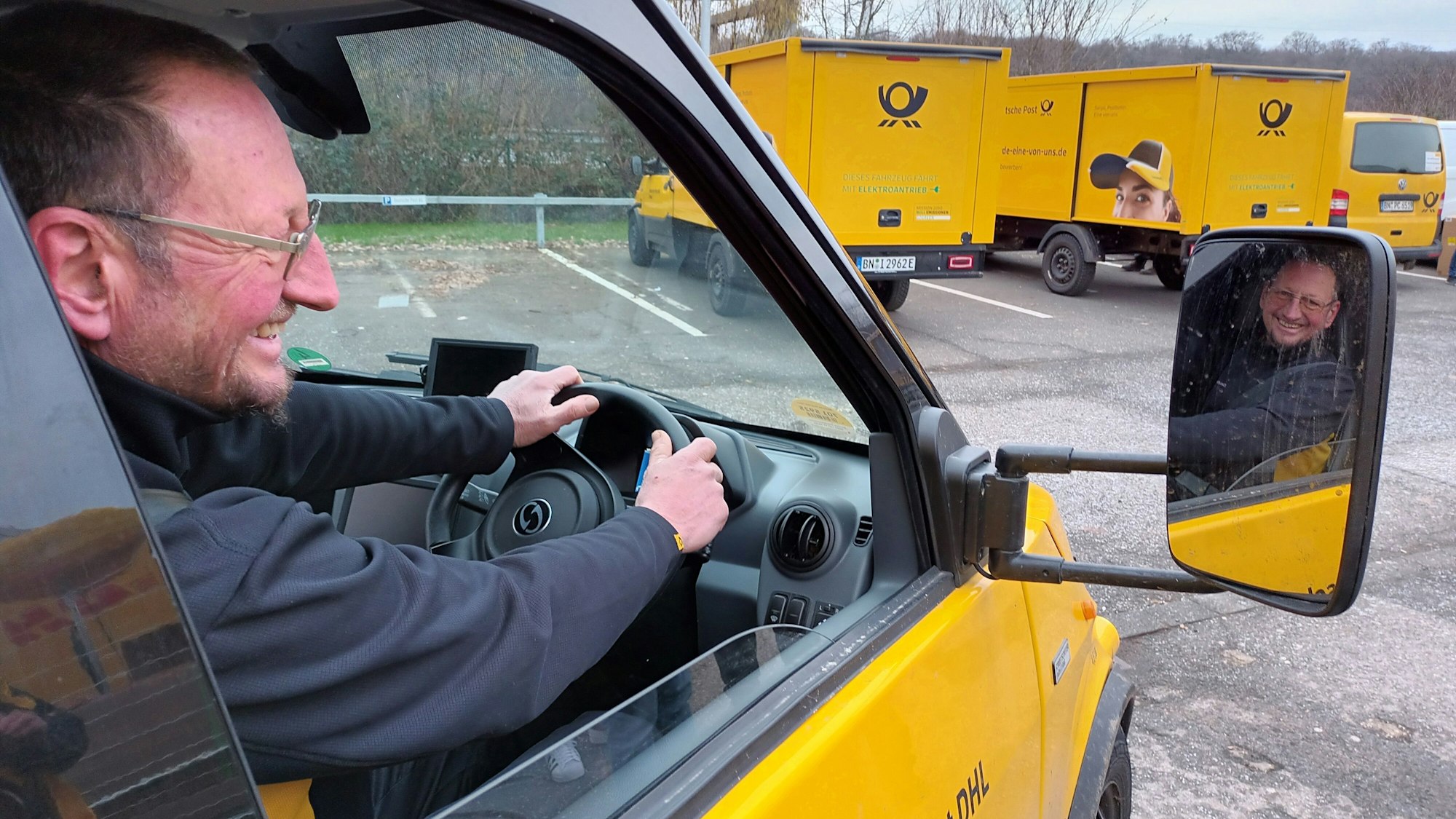 Ein Mann mit Brille sitzt am Steuer eines Zustellfahrzeugs der Deutschen Post, das auf der rechten Seite angebracht ist. Sein lachendes Gesicht spiegelt sich im Seitenspiegel.