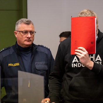 Man sieht drei Personen, die in der Mitte hält einen roten Hefter vor ihr Gesicht, die Personen links und rechts tragen Polizeiuniformen.