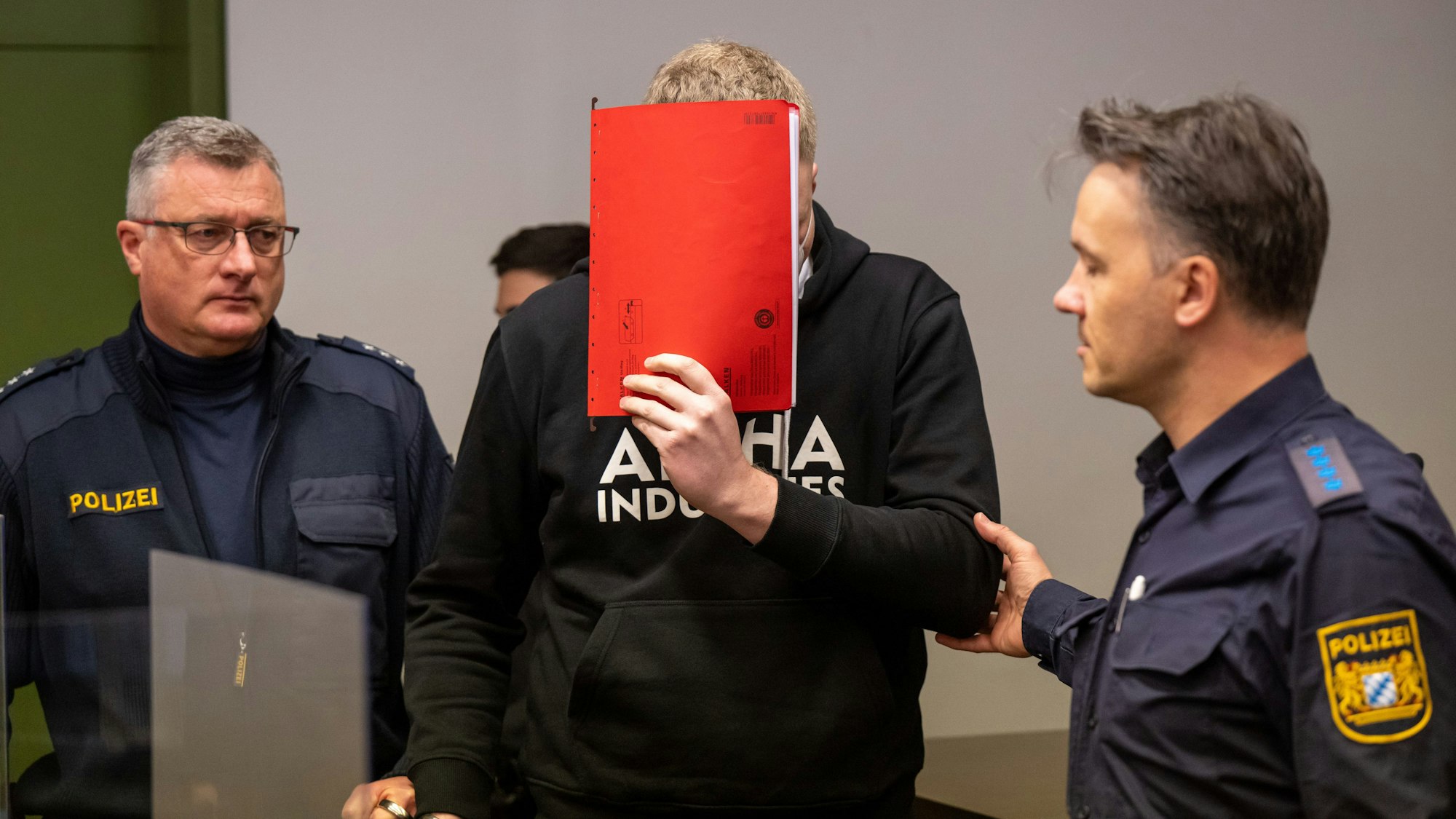 Man sieht drei Personen, die in der Mitte hält einen roten Hefter vor ihr Gesicht, die Personen links und rechts tragen Polizeiuniformen.